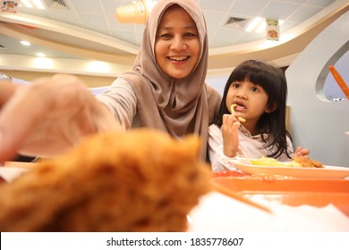 Asian Muslim Mother And Baby Girl Daughter Eating At Fast Food Restaurant, Family Enjoys Meals Of Fried Chicken, Burger And Potato Chips