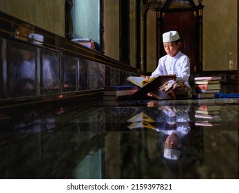 Asian Muslim Student Studying Al Quran Book To Understand Allah God Lesson In Islamic Mosque With Sttacked Of Al Quran Book Beside. Al Quran Book With Written Arabic Calligraphy Meaning Of Al Quran