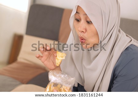 Asian muslim woman enjoys eating hot spicy casava or potato chips while sitting on sofa at living room, close up