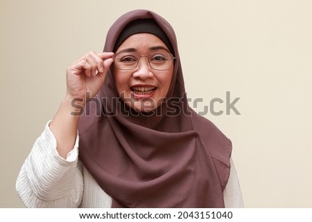 An Asian Muslim woman with grinning while holding her eye glasses, feeling awkward of something.
