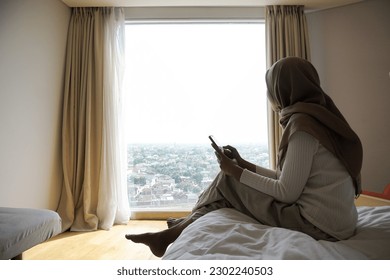 asian muslim woman sitting on bedroom bed in morning to enjoy outside view while using mobile phone - Powered by Shutterstock