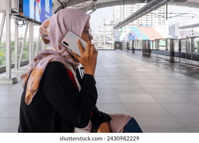 An Asian Muslim woman sitting on the bench at the train station and using her smart phone to call someone - Powered by Shutterstock