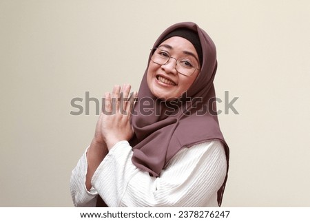 An Asian Muslim woman smiling and gives greeting hands with awkward expression.
