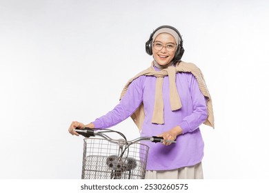 Asian Muslim woman walking with a bicycle while smiling broadly looking at the camera while using headphones on an isolated white background - Powered by Shutterstock