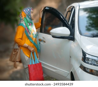 Asian Muslim woman wearing colorful hijab, orange shirt, carrying small red bag, opening white car door to go on holiday - Powered by Shutterstock