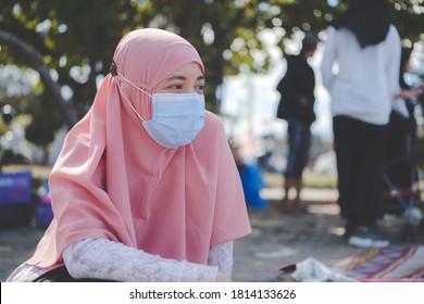 Asian Muslim Woman Wearing A Hijab And Surgical Mask Is Sitting On A Mat Outside. The Concept Of Preventing The Spread Of The Corona Virus Pandemic Or Covid-19