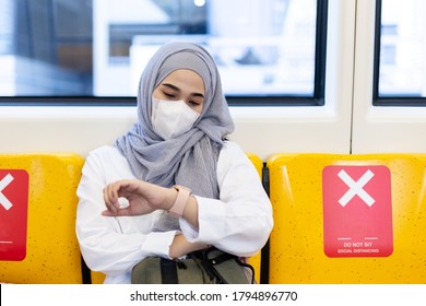 Asian Muslim Woman Wearing Medical Face Mask For Prevent Dust And Infection Virus Looking Wrist Watch Sitting On Chair With Symbol Social Distancing In Skytrain Transit System Public.