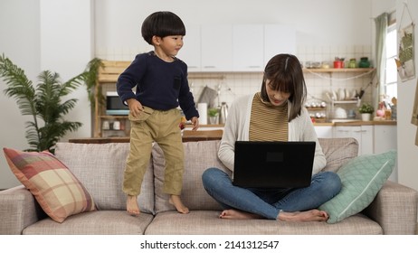 Asian Naughty Preschool Boy Bothering His WFH Mother With A Laptop In Living Room. She Shakes Her Head At Her Son While He Is Jumping Up And Down On Couch