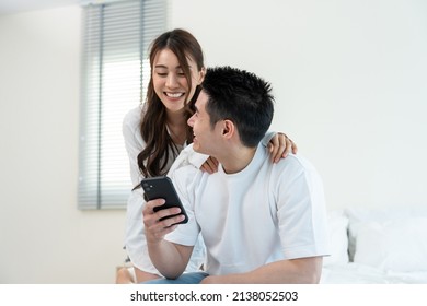 Asian New Marriage Couple Sit On Bed And Look At Telephone In Bedroom. Attractive Beautiful Young Man And Woman In Pajamas Enjoy Early Morning Activity And Holding Phone And Gossip Together At Home.