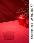 An Asian New Year table is decorated with a teapot, red envelopes, and a winterberry tree against a red backdrop, while there is a blank space for a products display. Template photo with a high angle.