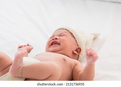 Asian Newborn baby crying sick fever lying on white bed. Adorable infant 0-1month fussy screaming unhappy angry. Child wearing pants and rabbit hat beige knitted tired and hungry.