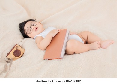 Asian Newborn Baby Wearing Glasses With Favorite Book On Beige Blanket , 3 Month-old Infant Lying In Bed With Relax. Adorable Baby Resting After Eat Full. Little Toddler Cute Sleeping Sweet Dreams.