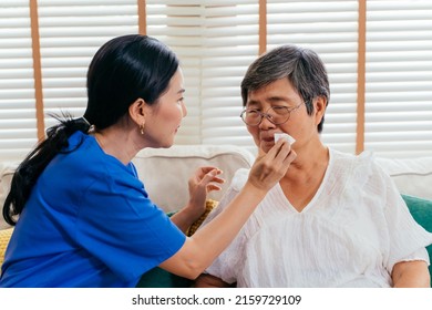 Asian Nurse In Blue Scrubs Wiping Lips Of Aged Woman With Tissue While Taking Care Of Patient On Couch At Home