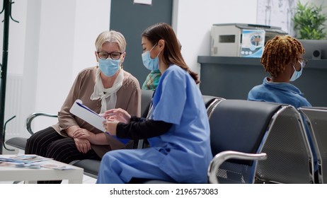 Asian Nurse Consulting Senior Woman With Face Mask In Hospital Reception Lobby, Talking About Disease Diagnosis And Healthcare. Specialist Giving Advice About Covid 19 Prevention. Handheld Shot.