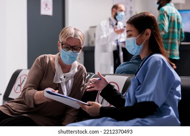 Asian Nurse Filling Out File Of Mature Adult Patient At Medical Clinic Waiting Room. Health Personnel Attending Patients At Hospital Emergency Room. Diverse People Wearing Masks In Doctor Office.