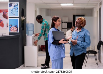 Asian Nurse Showing Disease Diagnosis To Person In Hospital Reception Lobby, Explaining Medical Results On Laptop. Diverse Women Talking About Medicine Treatment And Recovery In Waiting Area.