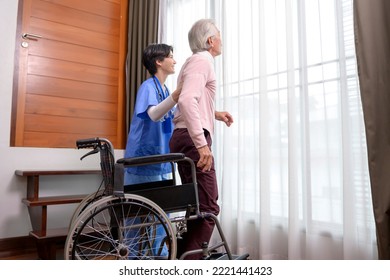 An Asian Nurse Taking Care Of An Elderly Man Getting Up From Wheelchair At Senior Healthcare Center.