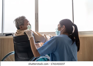 Asian nurse taking care of mature male patient sitting on wheelchair in hospital. Young woman and old man wearing surgical face mask for protection of covid 19 pandemic. Girl smile to elderly man.