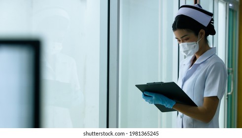 Asian Nurse Woman Checking Patient Chart As Routine, Nurse Look Through Windows Outside Care Unit Or Emergency Room In Hospital. Healthcare Hospitality Concept