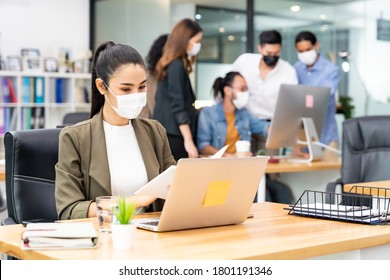 Asian Office Employee Businesswoman Wear Protective Face Mask Work In New Normal Office With Interracial Business Team In Background As Social Distance Practice Prevent Coronavirus COVID-19 Spreading.