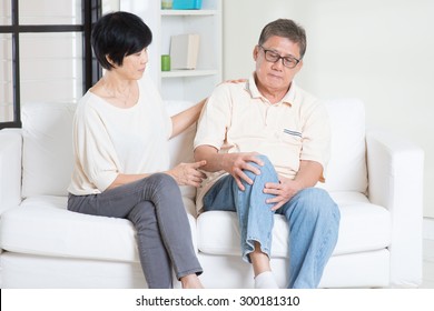 Asian Old Man Knee Pain, Sitting On Sofa With Wife At Home. Chinese Family, Senior Retiree Indoors Living Lifestyle.