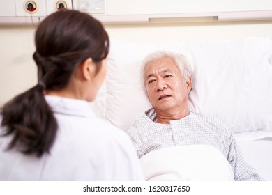 Asian Old Man Lying In Bed Talking To Young Female Doctor In Hospital Ward