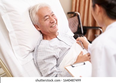 Asian Old Man Lying In Bed Talking To Young Female Doctor In Hospital Ward