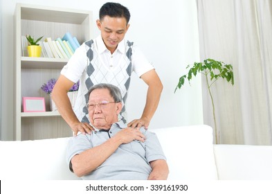 Asian Old Man Shoulder Pain, Sitting On Sofa , Son Massaging Father Shoulder. Chinese Family, Senior Retiree Indoors Living Lifestyle At Home.