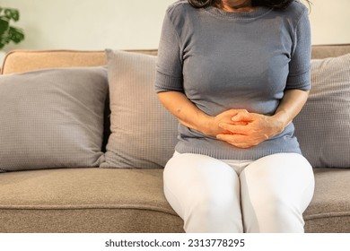 Asian old woman sitting on sofa with stomach ache irritable bowel syndrome Abdominal pain in the elderly, flatulence, inflammatory bowel disease, health care - Powered by Shutterstock