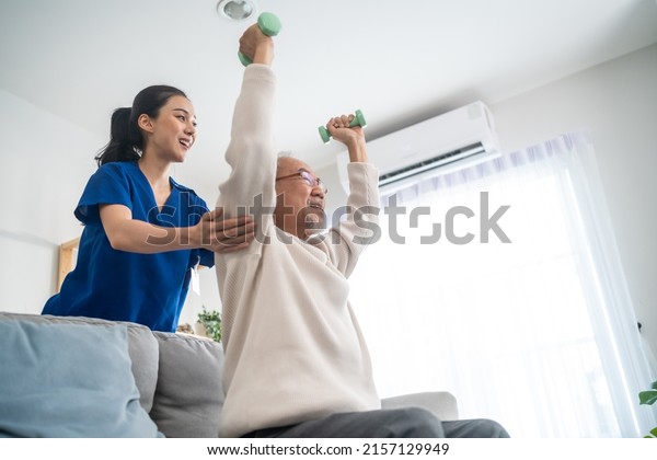 Asian older aged man doing physiotherapist with\
support from nurse. Senior elderly male sitting on sofa in living\
room using dumbbells workout exercise for patient with caregiver in\
nursing care.