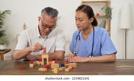 Asian Older Male Parkinson’s Patient Trying To Build Wood Brick With Hand Tremor While Having Rehabilitation At Home. Female Nursing Aide Giving Praise With Thumb Up Sign