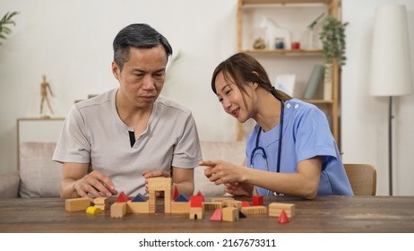 Asian Older Male Parkinson’s Patient Trying To Build Wood Brick With Hand Tremor While Having Rehabilitation At Home. Female Nursing Aide Giving Praise With Thumb Up Sign