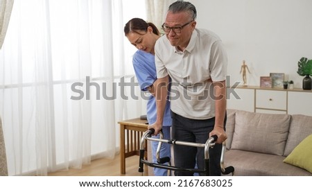asian older male stroke patient practicing using a walker with the assistance of his personal care attendant in the living room at home