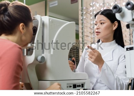 asian optometrist pointing with finger while measuring eyesight of blurred woman on vision screener