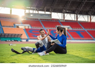 Asian para-athlete with prosthetic blades and trainer sit in stadium. Attractive amputee male runner and young sportswoman taking a break after practicing workout for Paralympics running competition - Powered by Shutterstock