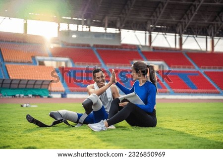 Asian para-athlete with prosthetic blades and trainer sit in stadium. Attractive amputee male runner and young sportswoman taking a break after practicing workout for Paralympics running competition