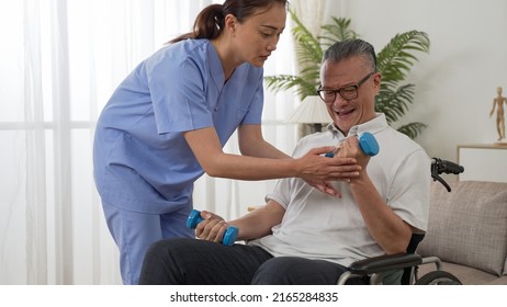 Asian Paralyzed Elderly Man Sitting In Wheelchair And Going Through Physical Therapy With Personal Care Attendant’s Support. He Does Weight Lifting Using Dumbbells At Home