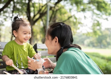 Asian Parent In Public Garden, Happy Baby Girl With Green T Shirt Eating Yogurt Lookat Her Mom, Smiling