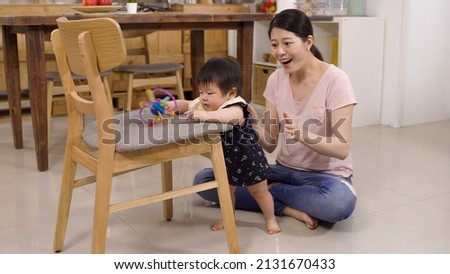 asian parent seated on the floor at home is clapping hands happily after letting go of her kid to stand by leaning on a chair with a toy.