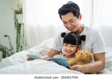 Asian Parent Spend Leisure Time With Kid Daughter In Bedroom At House. Happy Family, Loving Father Teach Young Preschool Little Kid Girl To Read Book. Parenting Relationship Home Education Concept.