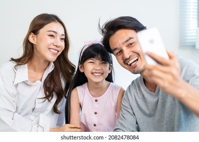 Asian Parents With Little Kid Daughter Video Call Online With Family. Loving Father, Mother And Young Girl Sitting On Bed In Bedroom Enjoy Using Mobile Phone Talking To Relative On Weekend In House.