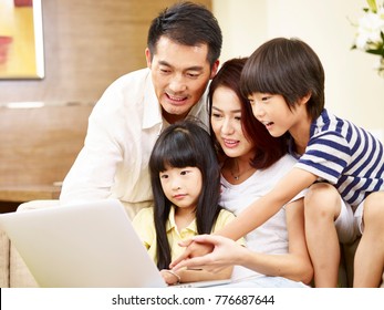 Asian Parents Mother And Father And Two Children Son And Daughter Sitting On Family Couch Using Laptop Computer Together.