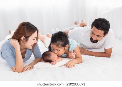 Asian Parents And Newborn With Sister Spend Time Together In Bedroom. Sister Kiss Adorable Infant While Baby Sleeping Among Happy Family, Mother And Father Lying In Bed With Two Daughters.