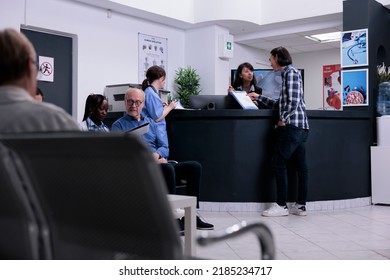 Asian Patient Handing Clipboard With Completed Form To Hospital Receptionist Standing At Front Desk. Man Attending Doctor Appointment In Busy Private Clinic With Diverse Patients.