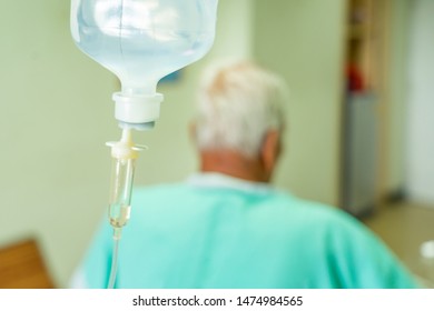 Asian Patient Older Man Lying At Hospital Bed With Saline Drip Showing Victory Sign. Drop Of Saline Solution To Help Patient, Iv Fluid Use For Intravenous Volume. Selective Focus