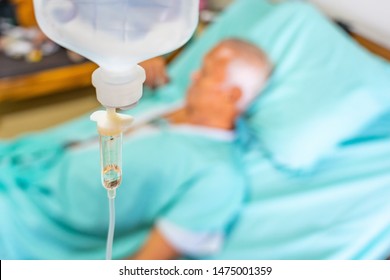 Asian Patient Older Man Lying At Hospital Bed With Saline Drip Showing Victory Sign. Drop Of Saline Solution To Help Patient, Iv Fluid Use For Intravenous Volume. Selective Focus