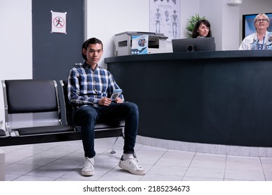 Asian Patient Sitting Down In Hospital Lobby Waiting For Doctor In Private Clinic To Perform Clinical Examination. Young Man At Hospital Reception Waiting For Receptionist To Call Him For