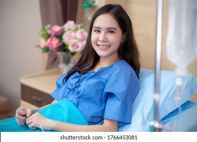 Asian Patient Woman Sick Saline Intake In Hospital. Beautiful Lady With Patient Dress, Smile Get Well Sitting On Patient's Bed In Hospital. Saline Bottle Hanging, Flower Vase Are At Room