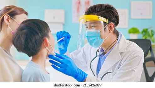 Asian Pediatrician With Face Shield And Mask Use Tongue Depressor To Exam Sick Boy Mouth When Mother Sit By In Clinic