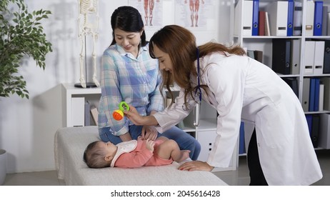 Asian Pediatrician Stooping Over The Baby On Treatment Couch. Observing Its Response To The Rattle Sounds. Well-child Exam. Newborn Hearing Screening Test Concept. Genuine Lifestyle